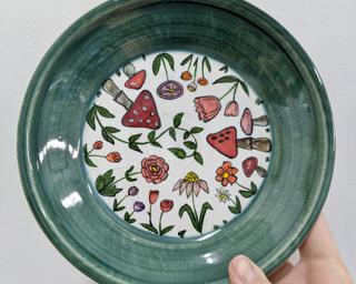 This wheel thrown dish is made with a white stoneware clay and has been carefully decorated with various flowers, leaves, and mushrooms. Some pinholing