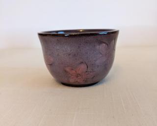 A small handle-less ceramic cup with a subtle purple glaze over a mid-fire dark brown clay painted with flowers.